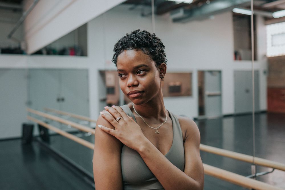 Person in a dance studio practicing ballet poses near a barre in black attire.