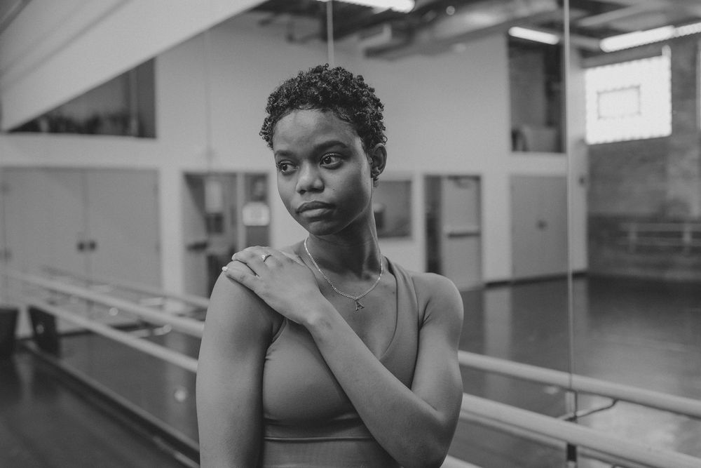Person in a dance studio practicing ballet poses near a barre in black attire.
