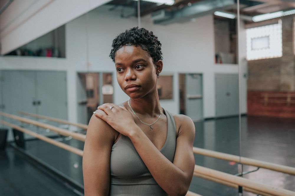 Person in a dance studio practicing ballet poses near a barre in black attire.
