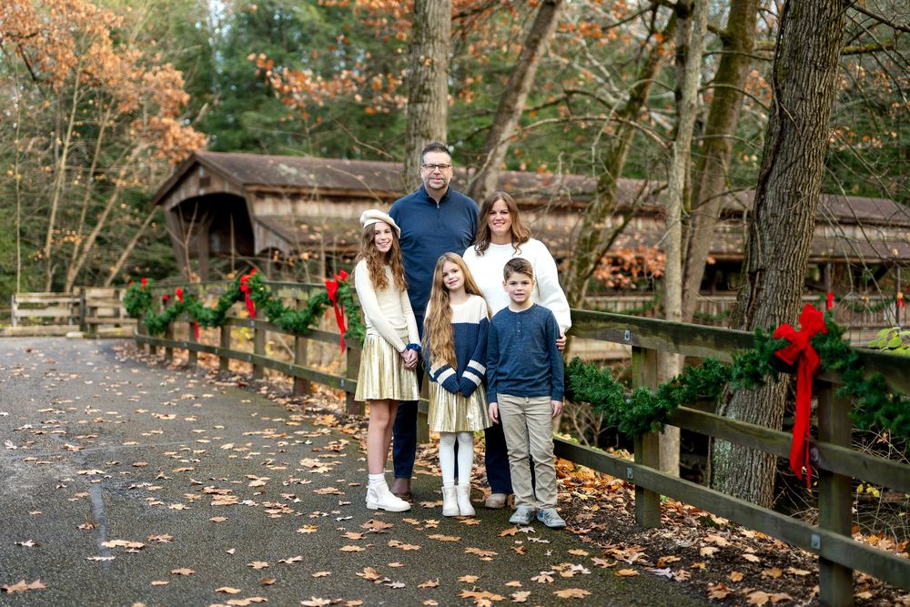Family of five posing on a leaf-covered path by a Christmas-decorated covered bridge in autumn woods.