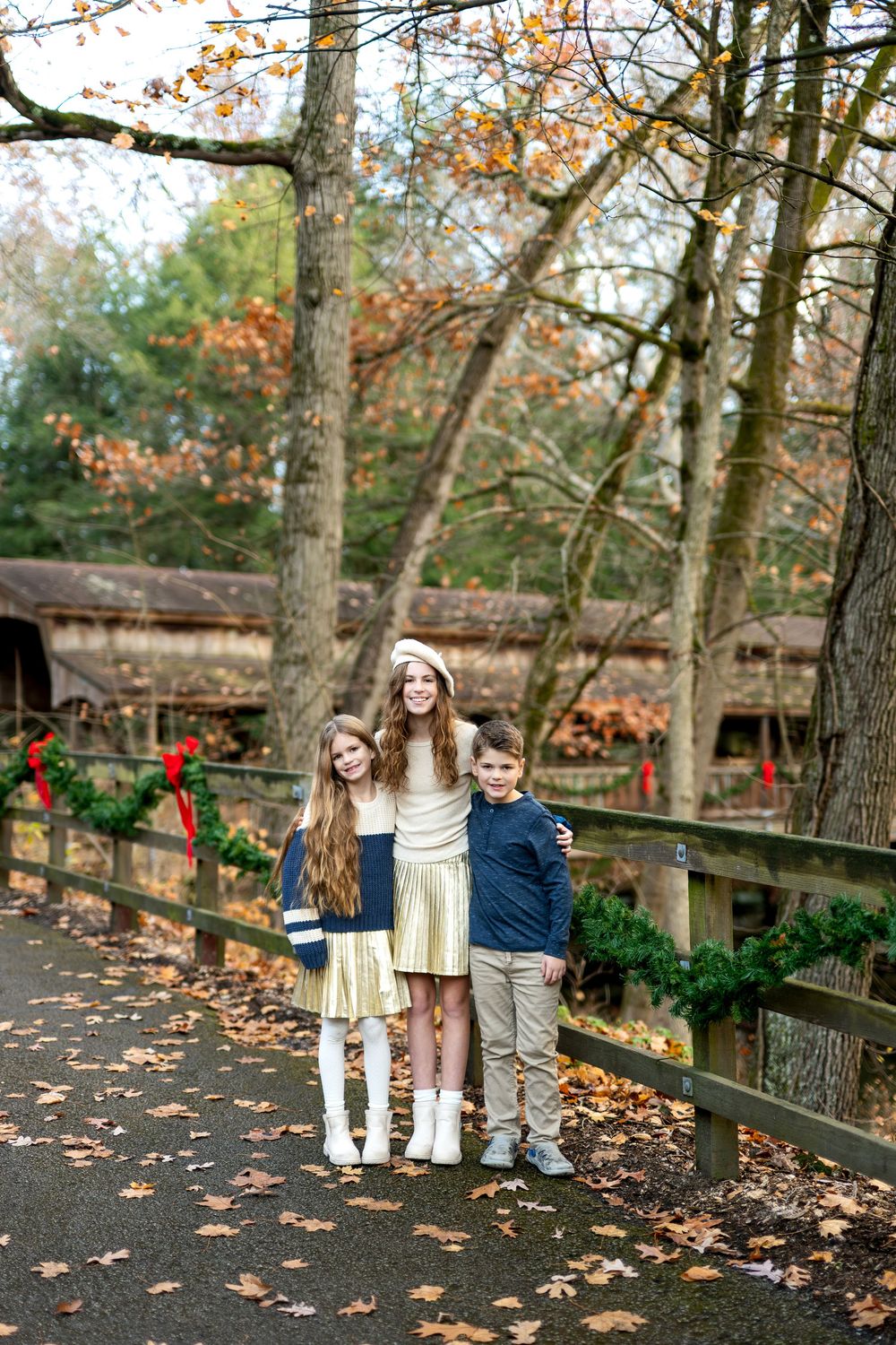 Three siblings hugging and smiling on a fall path beside a garland-decorated covered bridge.