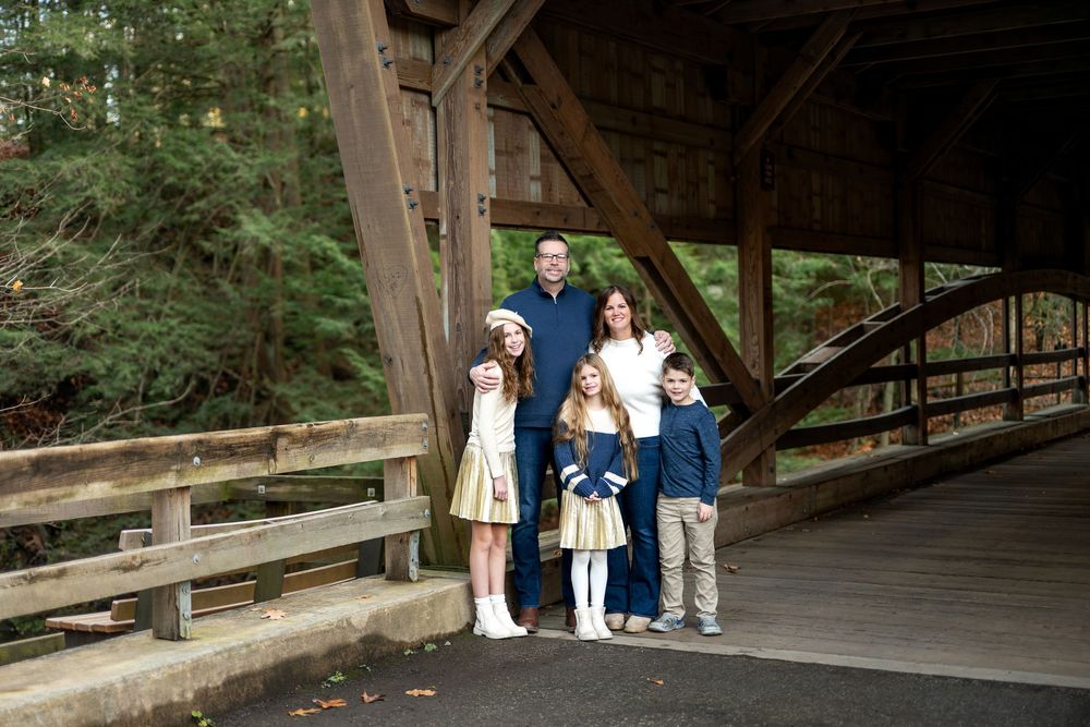 Family of five posing together at the entrance of a rustic wooden covered bridge in the forest.