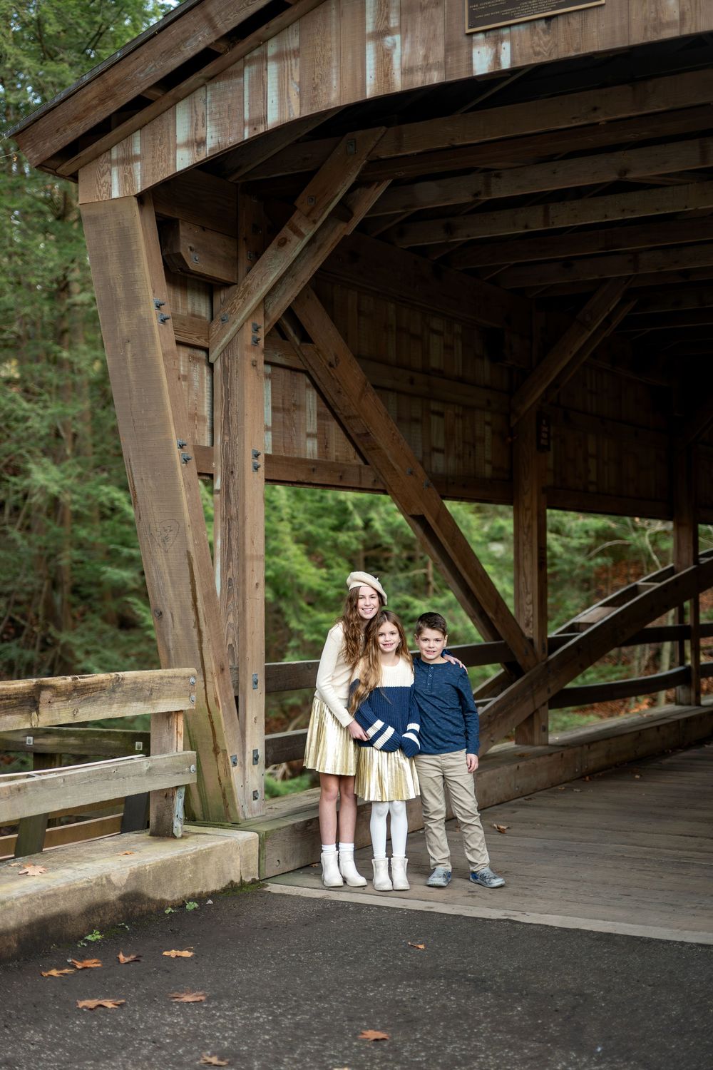 Three children standing together under the wooden structure of a rustic covered bridge in the forest.