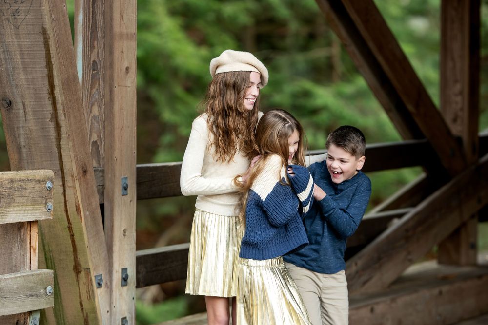 Three siblings laughing and playing together under the wooden beams of a covered bridge.
