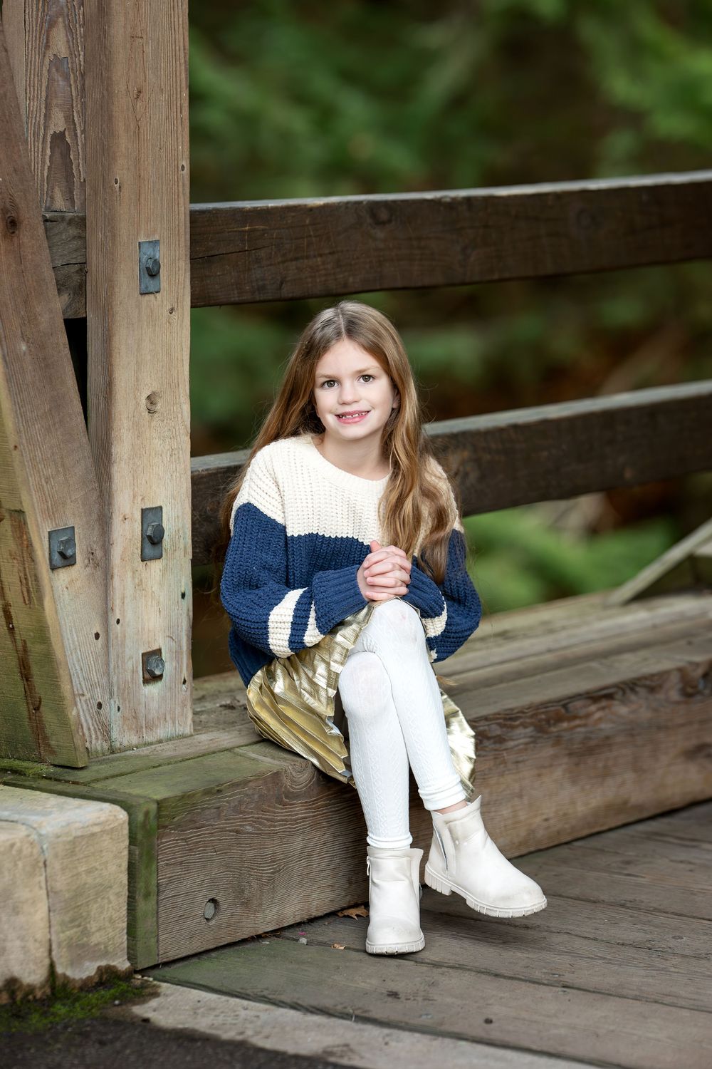 Young girl in a navy and cream sweater with gold skirt sitting on a wooden bridge ledge.