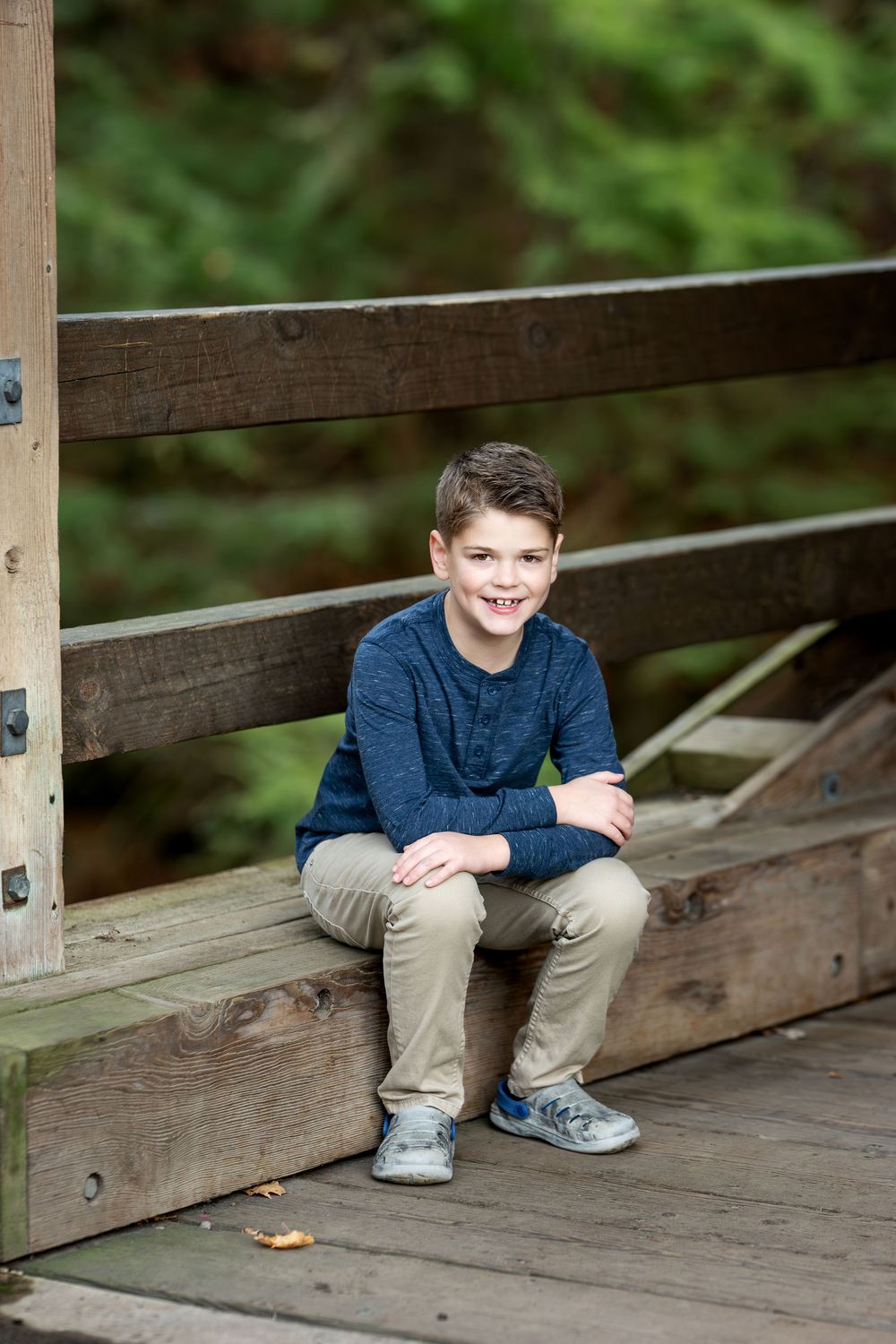 Smiling young boy in a navy shirt and khaki pants sitting on a rustic wooden bridge.