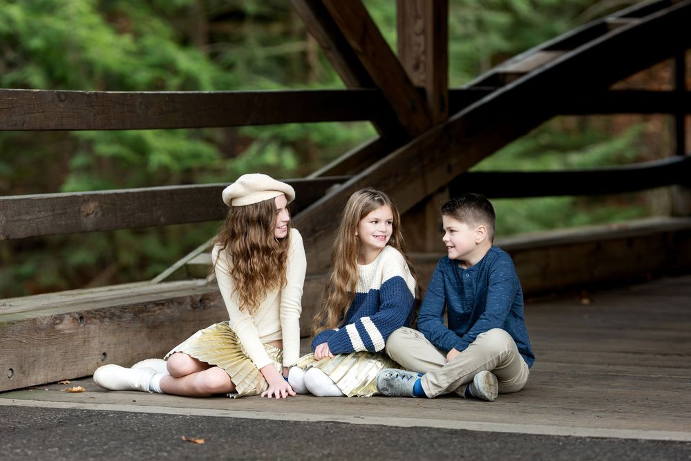 Three children sitting together on the floor of a rustic wooden covered bridge in the forest.