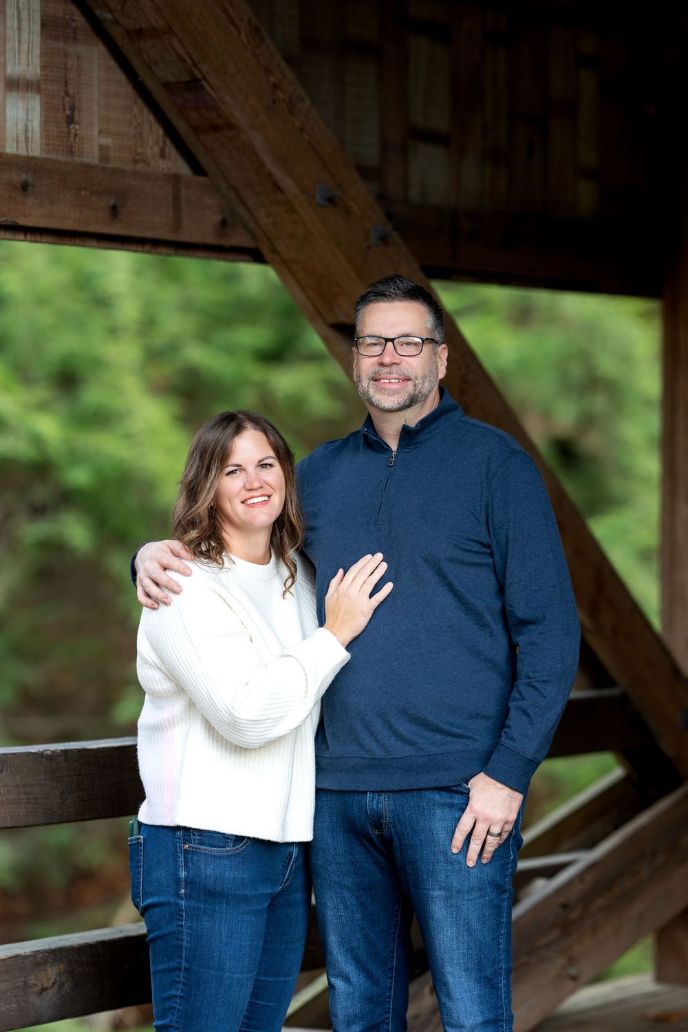 Couple smiling together under wooden bridge structure with lush green trees in background.