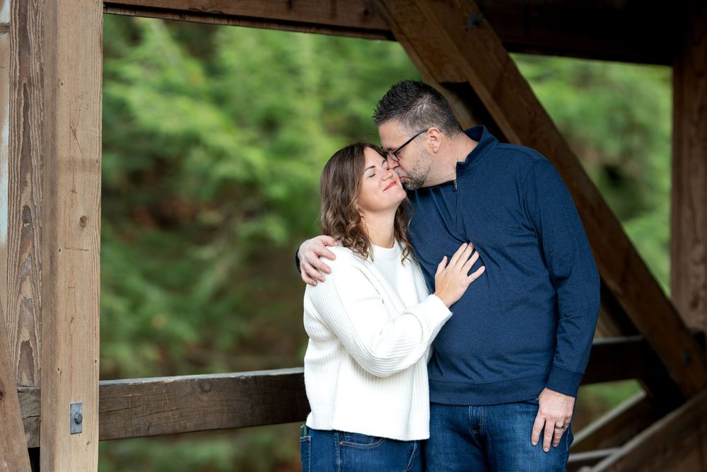 Man kissing woman on cheek as she smiles under rustic wooden covered bridge.