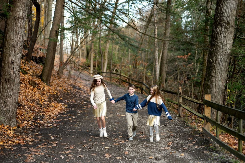 Three children holding hands walking along a wooded forest trail in autumn.
