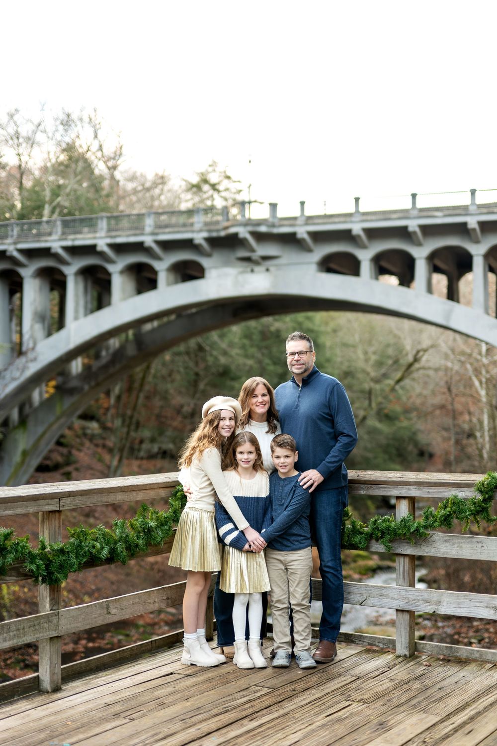 Family of five posing on wooden deck with arched stone bridge backdrop.