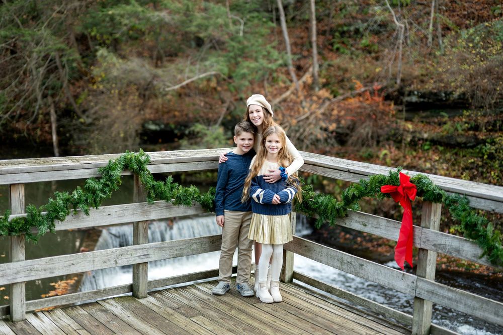 Three siblings hugging on a holiday-decorated wooden bridge with waterfall in background.