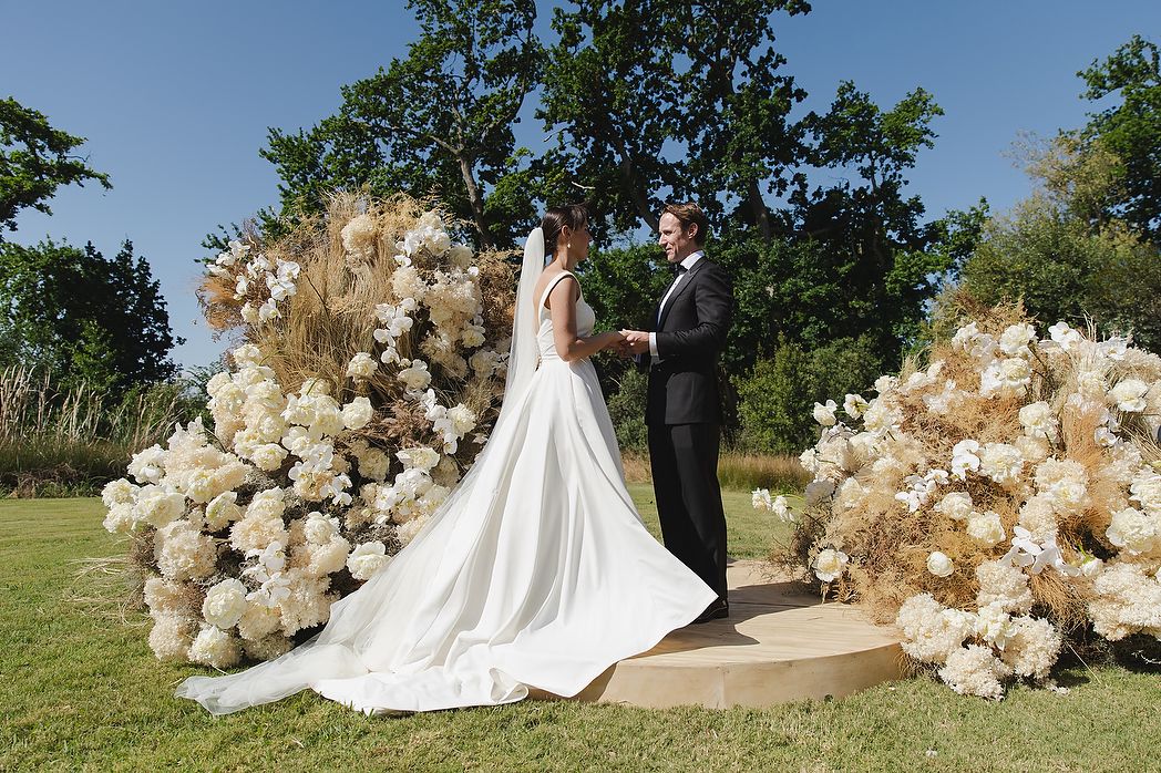 The couple holding hands during the ceremony with sculptural florals in the background.