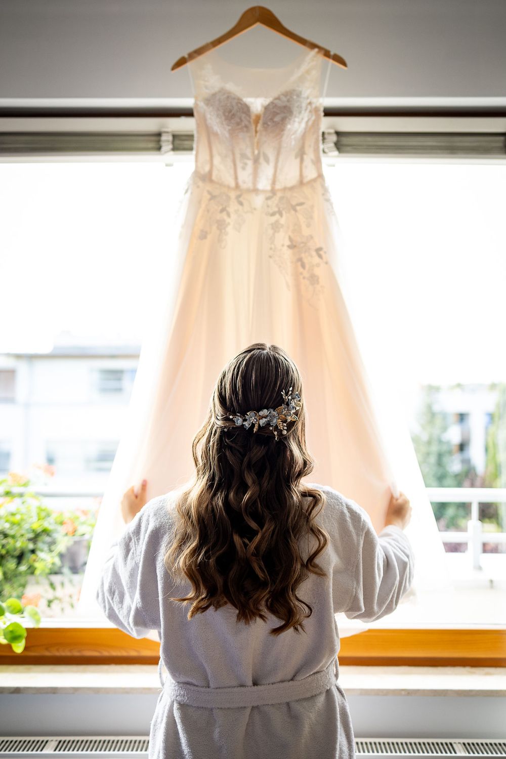 A wedding dress hangs in front of a window while someone in a white robe gazes up at it.
