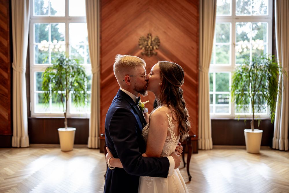 A romantic moment between newlyweds in an elegant room with large windows and potted plants.