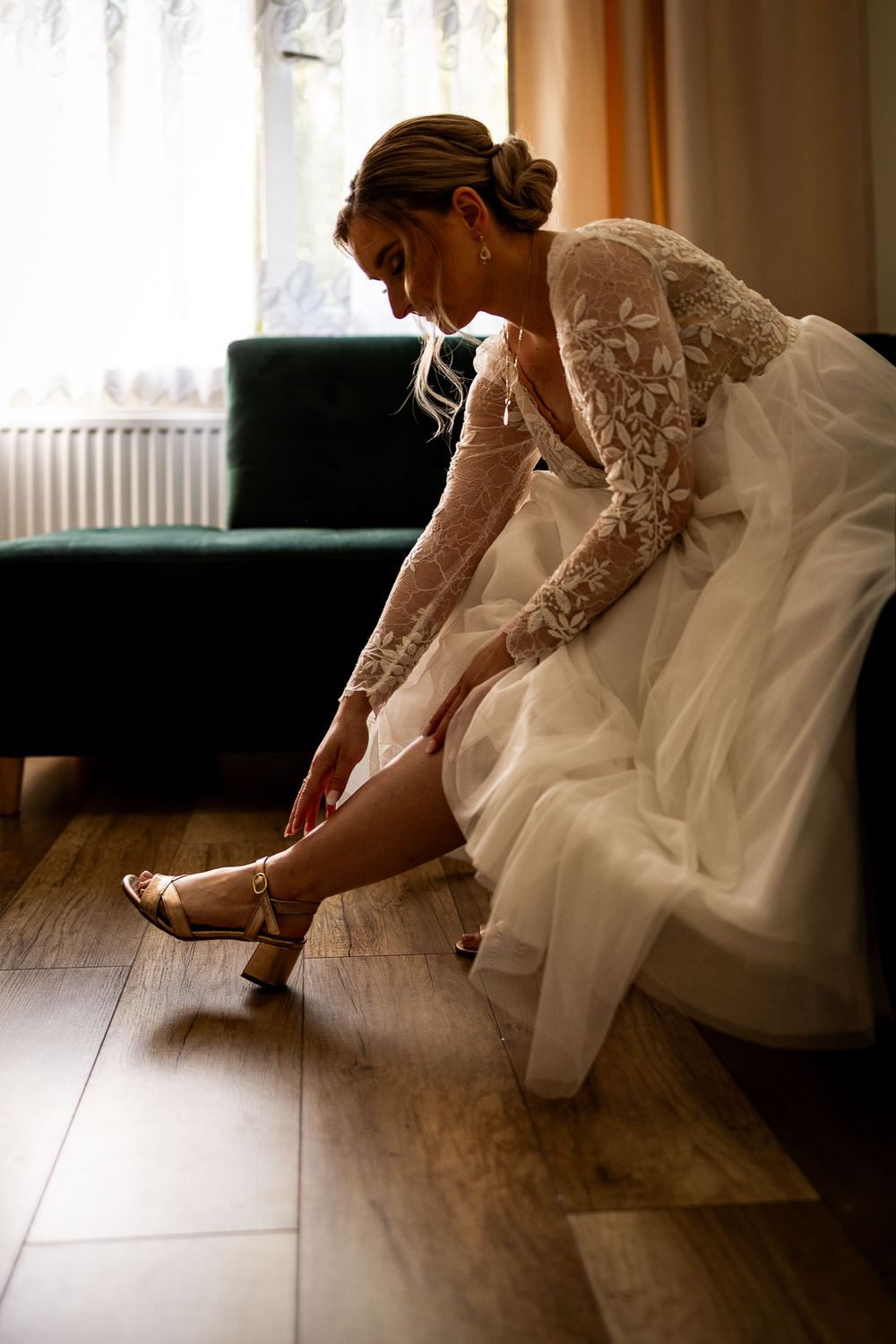 A bride in a lace wedding dress putting on her shoes while seated on a green couch.