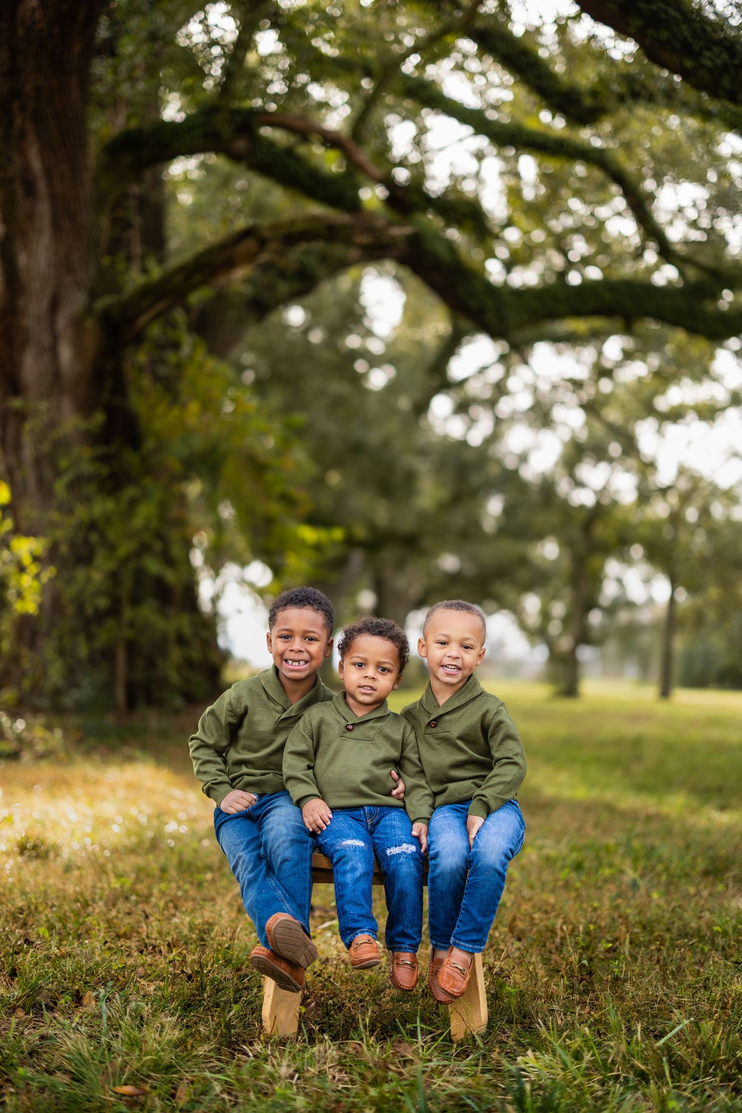 Three children in matching olive green shirts sitting together in grass under large trees.