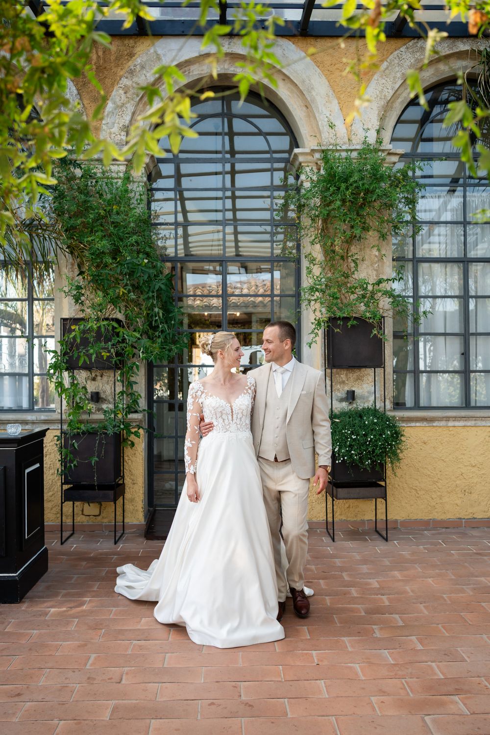 A romantic moment captured in an elegant indoor venue with terracotta floors and arched windows adorned with greenery.