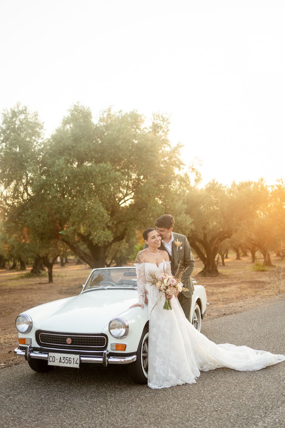 A couple poses with a classic white Fiat convertible in an olive grove during sunset.