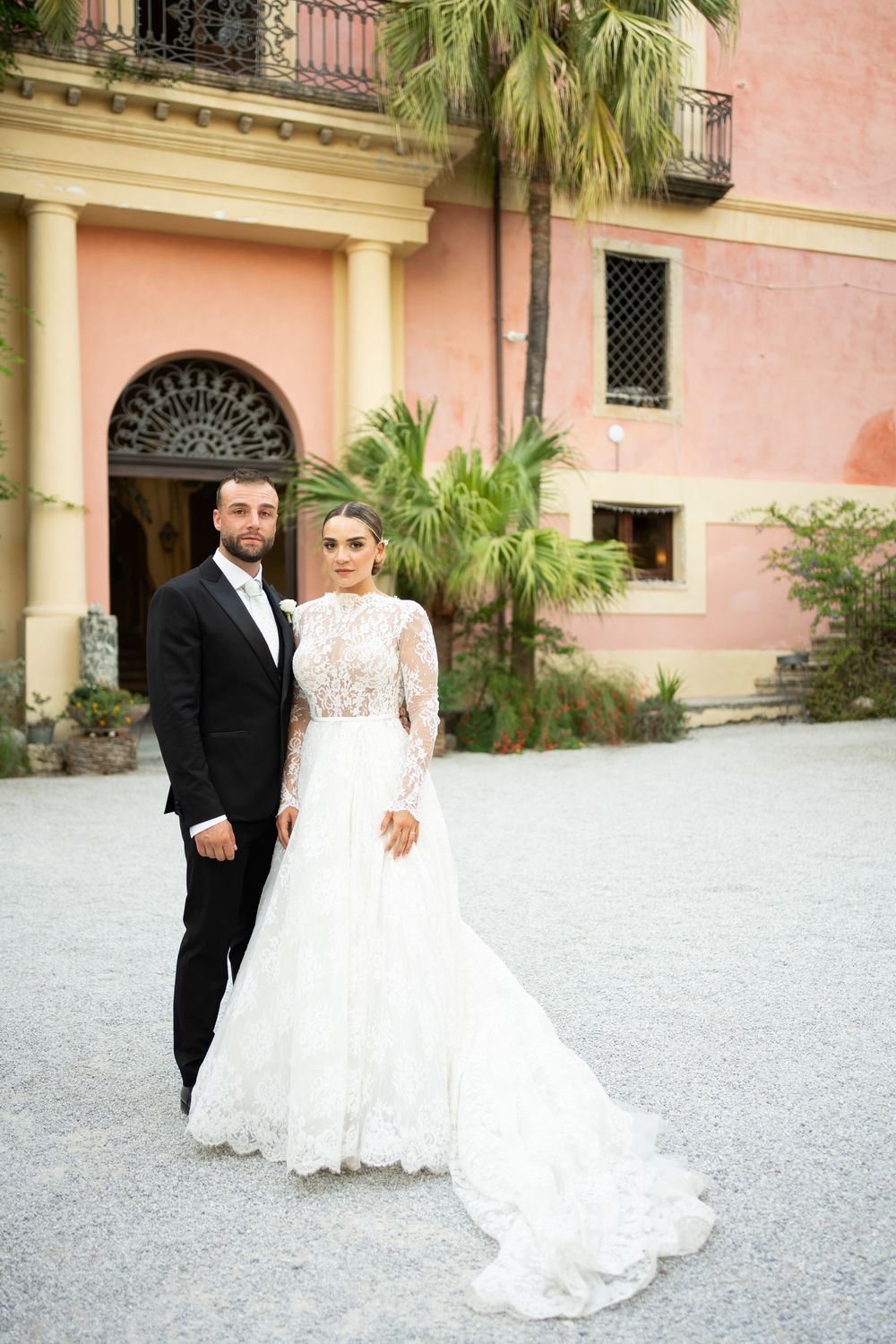 A couple poses outside a pink Mediterranean-style building with palm trees and elegant columns.