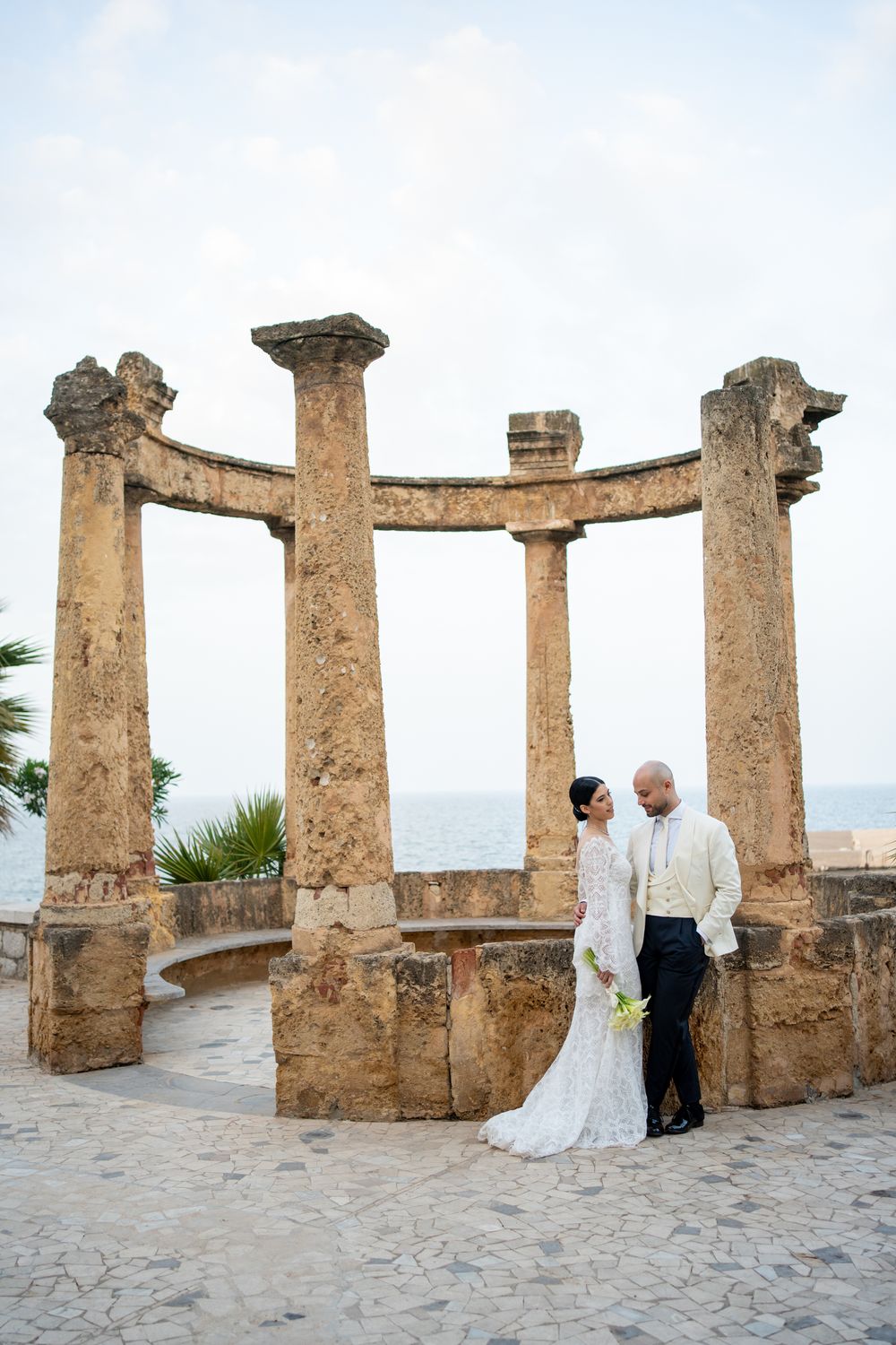 Ancient stone columns form a curved semicircle overlooking the ocean at a scenic Mediterranean coastal viewpoint.
