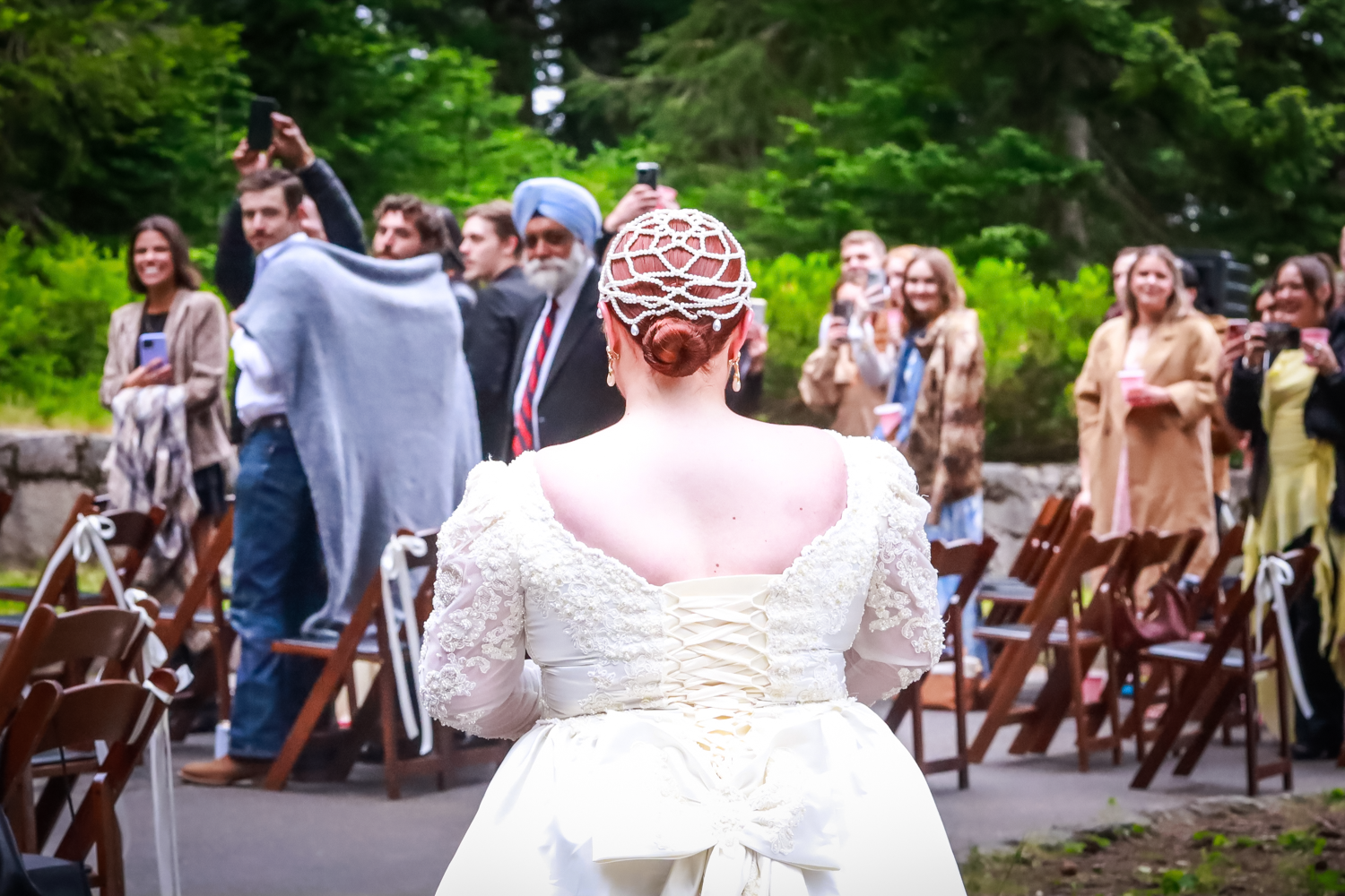 A wedding gathering in a lush outdoor setting with wooden chairs and guests mingling around a person in white dress.
