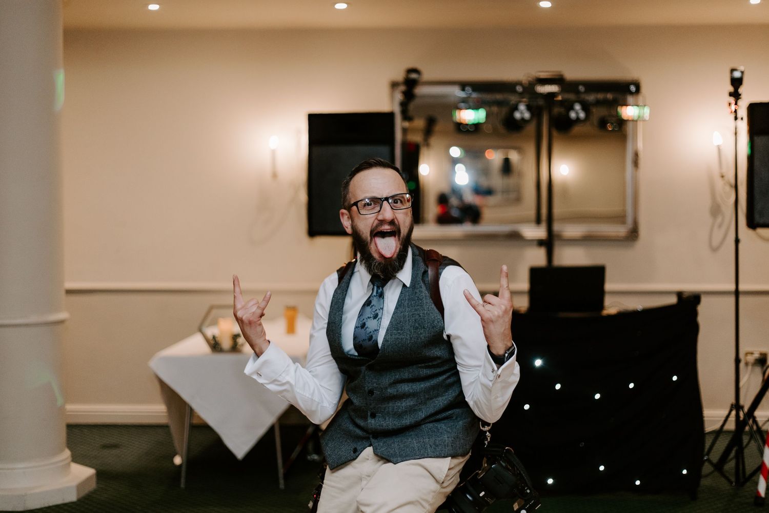 A person in a vest and tie celebrates enthusiastically at an indoor event venue with lighting equipment visible.