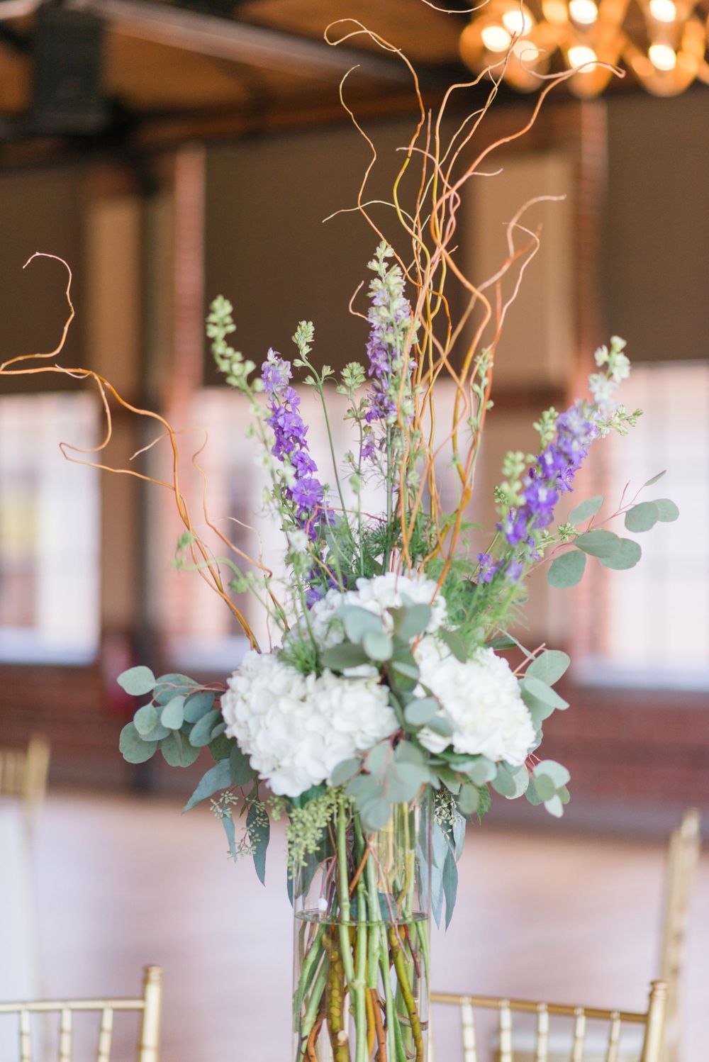 Elegant wedding centerpiece with purple delphinium, white hydrangeas, and curly willow branches in a tall glass vase.