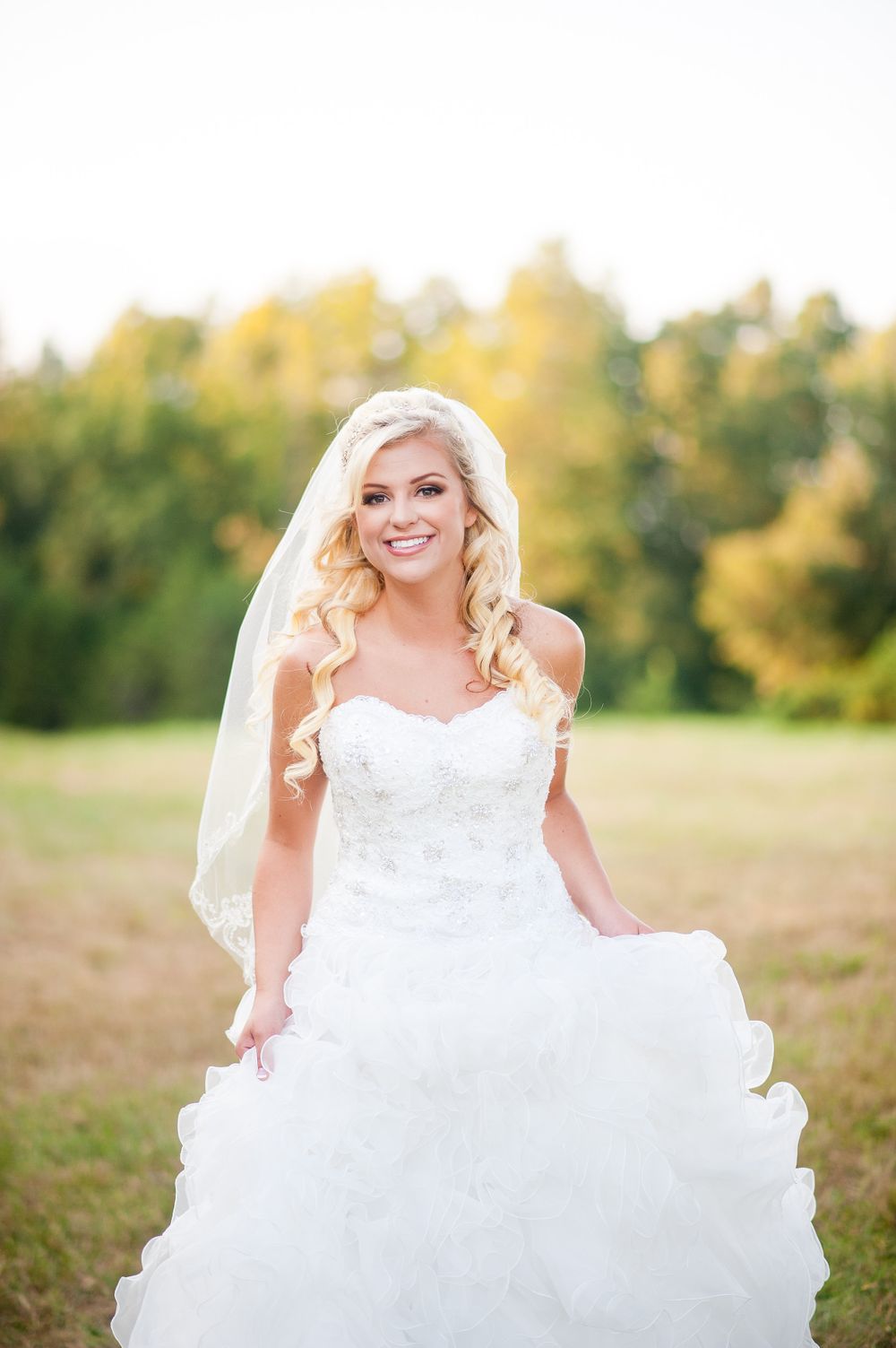 Bride in a ruffled ballgown poses in a sunlit field with autumn trees in the background.