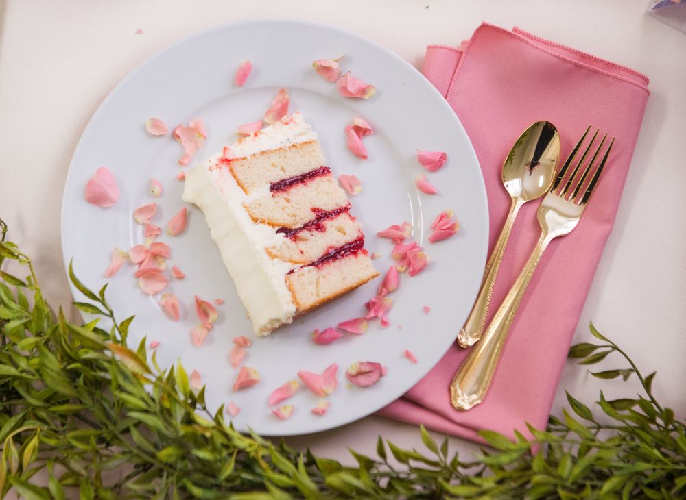 Slice of white wedding cake with pink rose petals on blue plate with gold utensils.