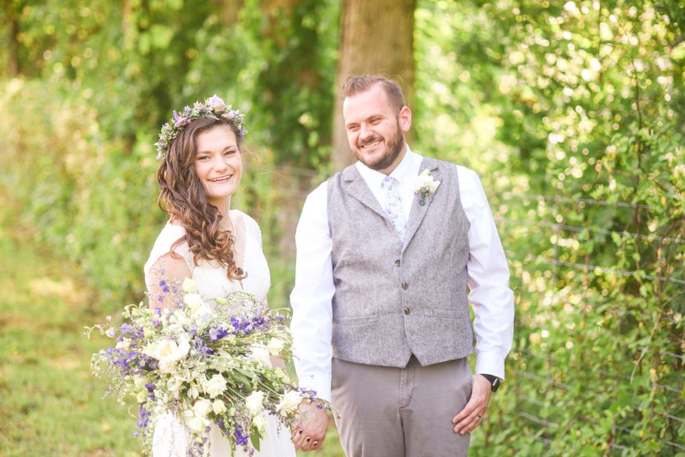 A couple shares a joyful moment in a sunlit garden setting with the bride holding a purple wildflower bouquet.