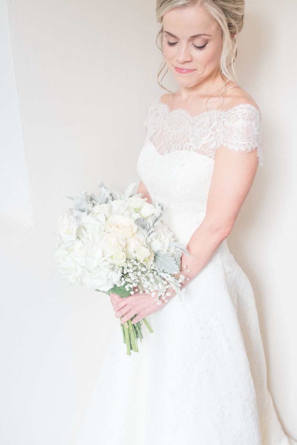 A bride in an off-shoulder lace gown holds a white and silver bouquet against a light background.