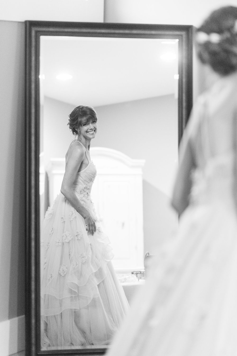 Black and white reflection of a bride in a flowing wedding dress looking at herself in a mirror.