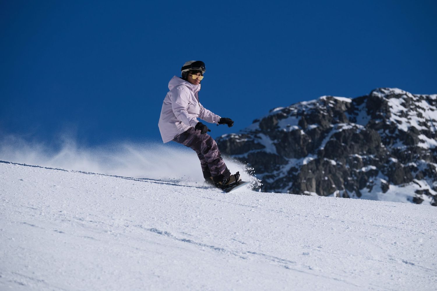 A snowboarder performs aerial tricks against a snowy mountain backdrop on a bright winter day.