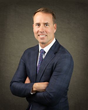 Professional headshot portrait of someone in a navy suit and purple patterned tie against a dark gray backdrop.