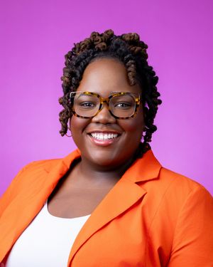Professional headshot against a pink background showing bright orange blazer, glasses, and curly hairstyle with a warm smile.