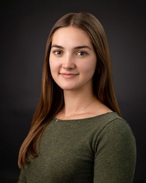 A person in a dark olive green sweater poses against a dark background in a professional headshot series.