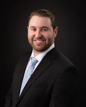 Professional headshot portrait of someone in a black suit and light blue tie against a dark background.