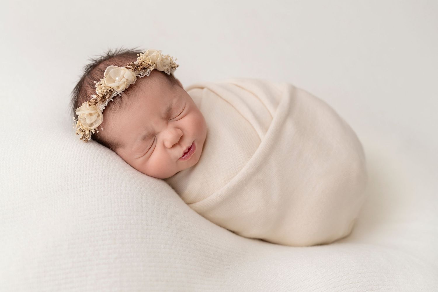 A newborn baby wrapped in a cream colored swaddle blanket and wearing a floral headband lays peacefully on a white blanket.