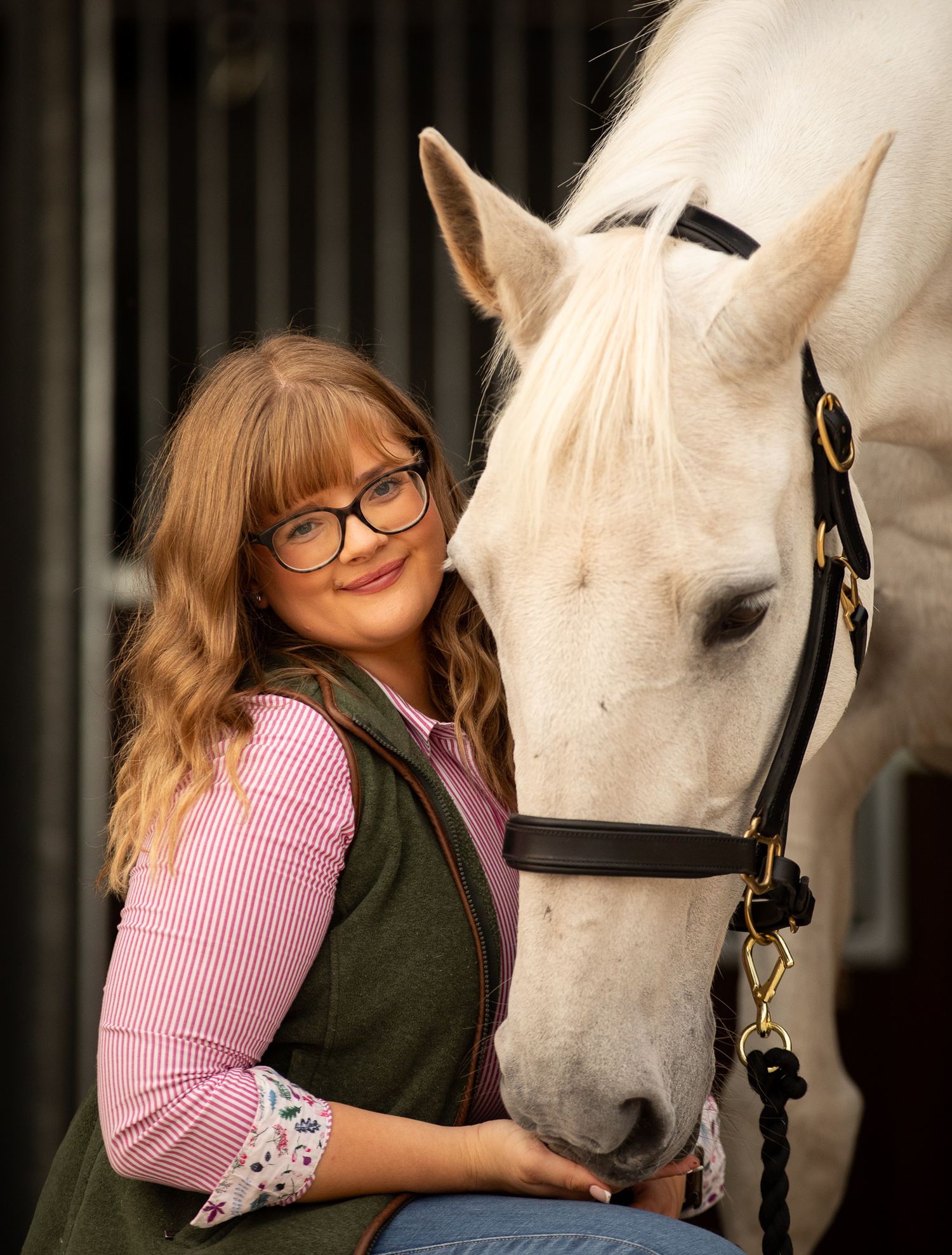 A young rider in a blue polka dot dress poses affectionately with a chestnut brown horse in an outdoor setting.