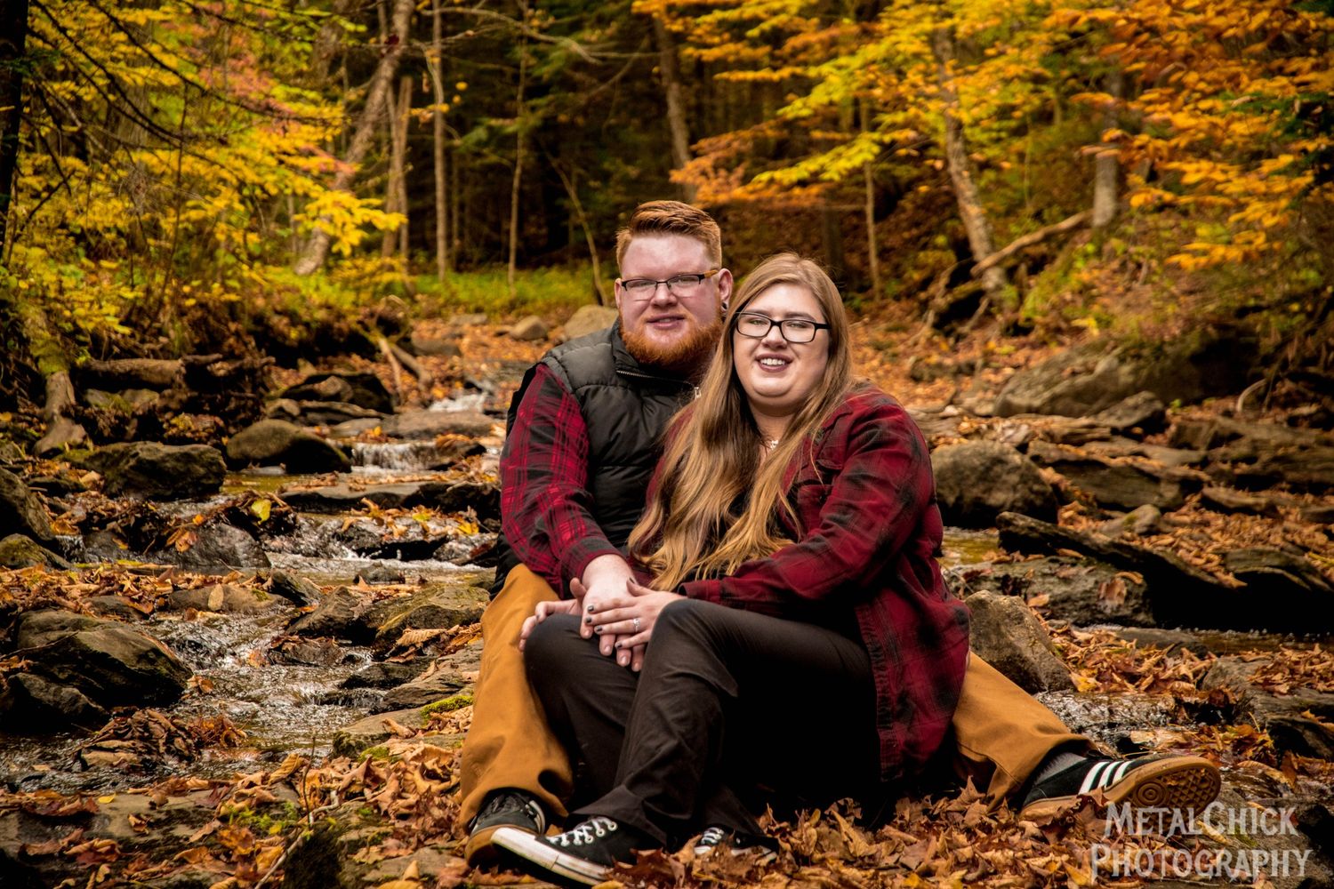 Engagement portrait by Northern Exposure featuring a couple sitting on some rocks in the middle of a stream in fall.