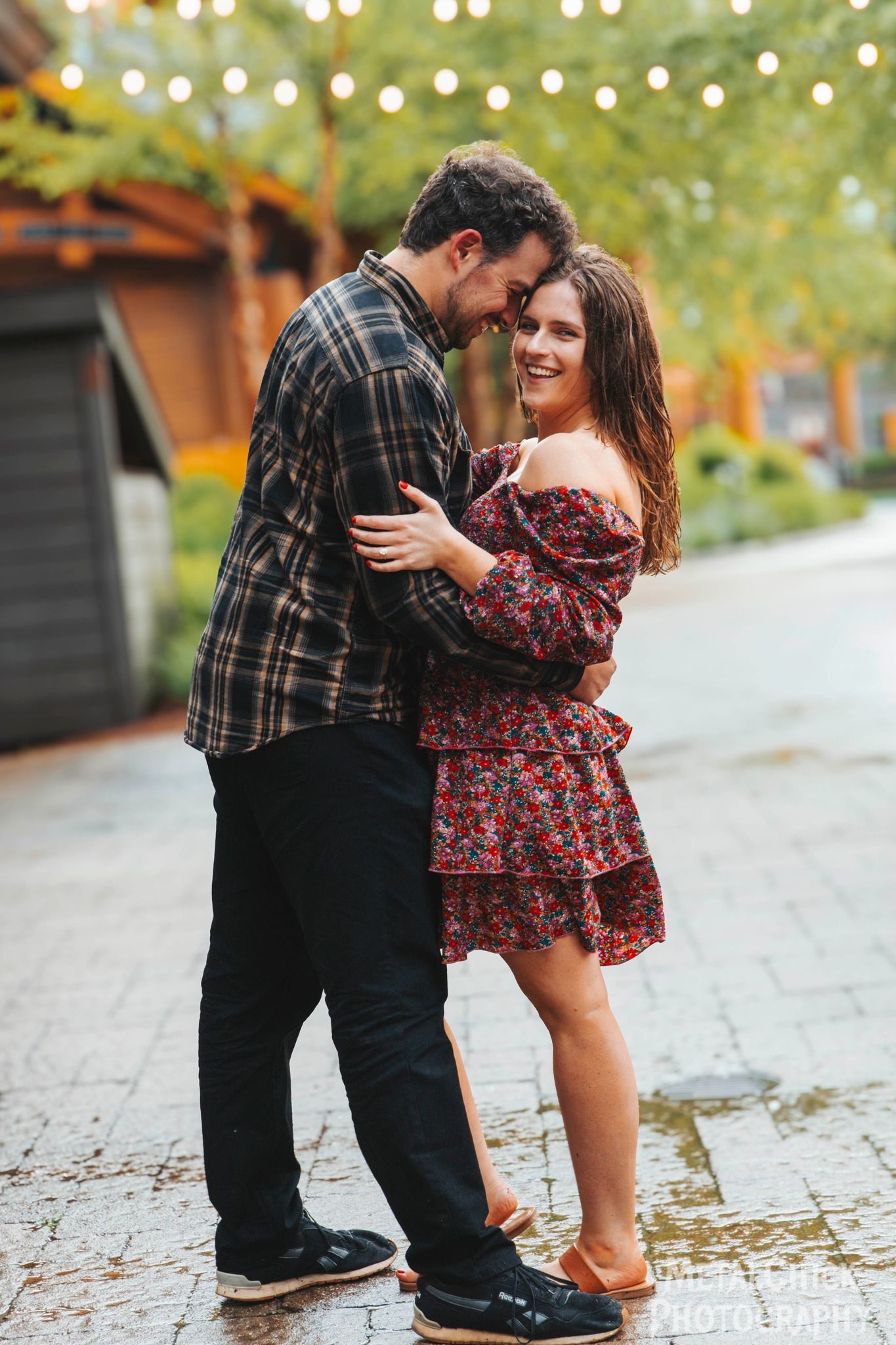 Engagement portrait by Northern Exposure: A smiling woman in a floral dress and a man in a flannel shirt embracing on a wet cobblestone stre