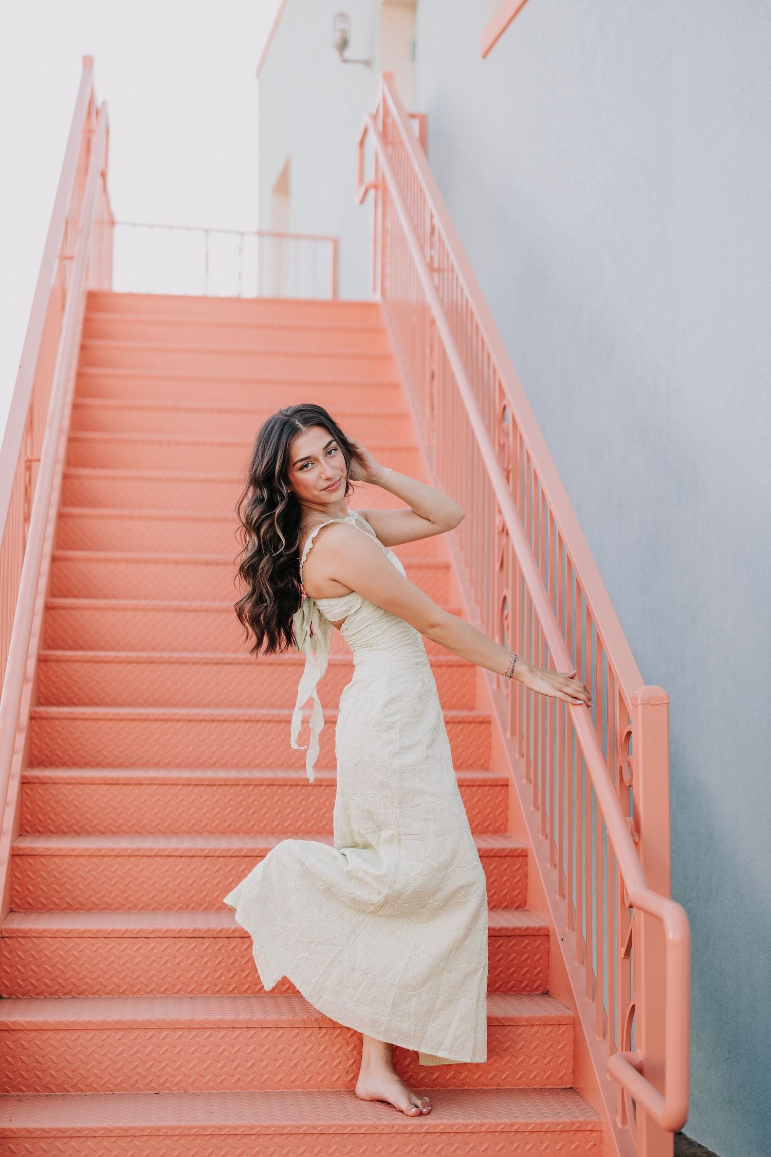Person in white dress posing on coral-colored staircase against light blue wall.