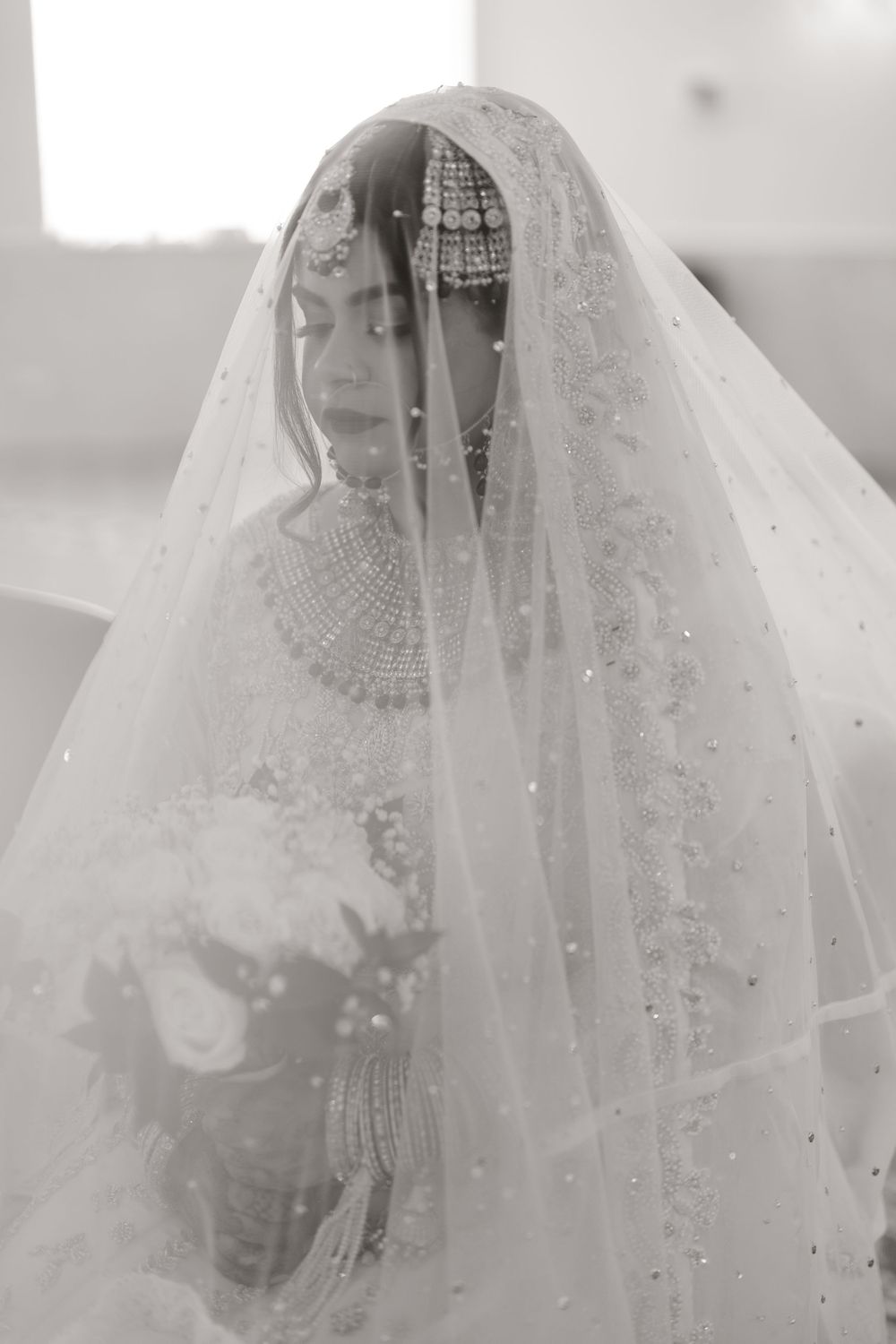 Elegant Muslim bride draped in embellished white veil holds bouquet in serene black and white wedding portrait.