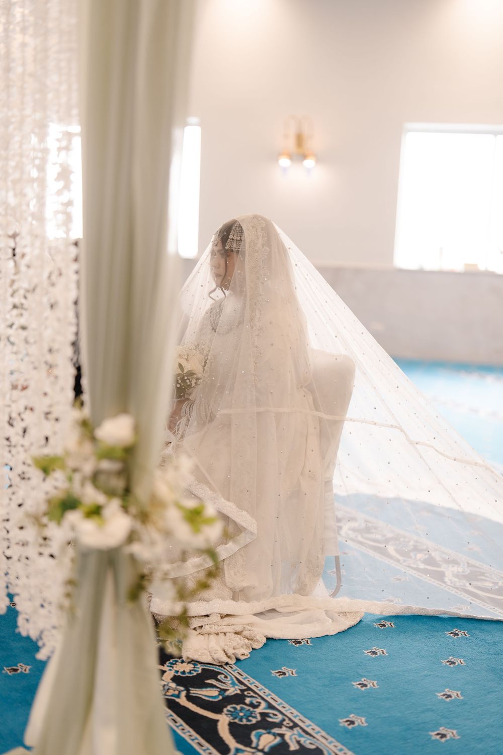 Muslim bride in ornate white wedding dress and veil kneels in prayer on blue carpet in mosque.