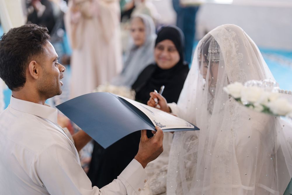 Groom signing nikah marriage contract while bride in white lace hijab watches during Islamic wedding ceremony.