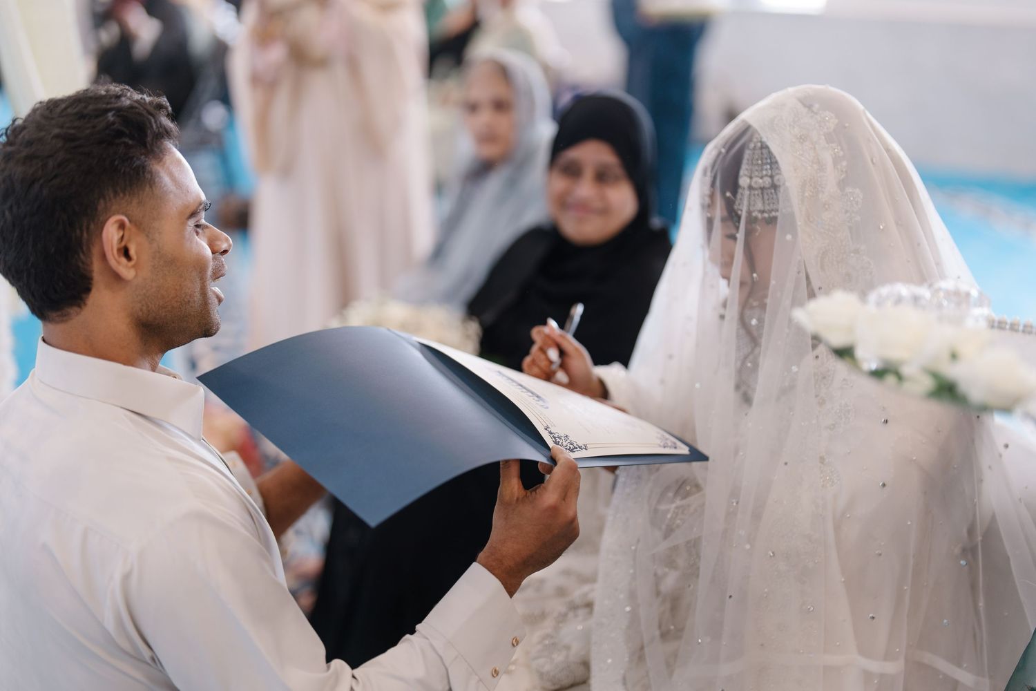 Groom signing nikah marriage contract while bride in white lace hijab watches during Islamic wedding ceremony.