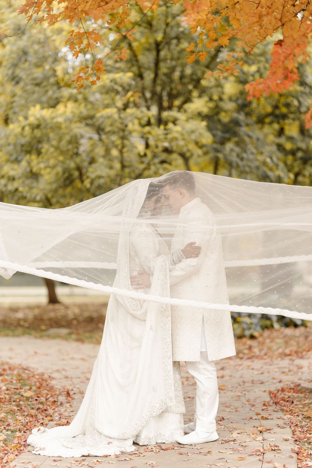 Muslim wedding couple embrace under flowing white veil surrounded by colorful autumn trees in park.