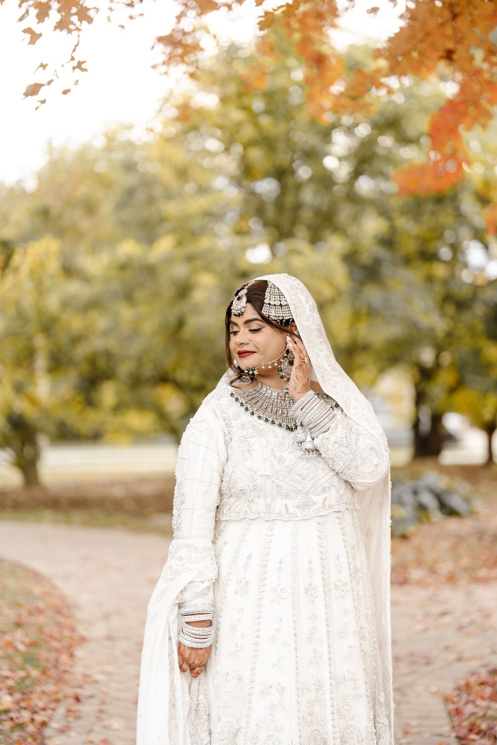 Beautiful South Asian bride in white lehenga with silver jewelry poses gracefully in autumn park setting.