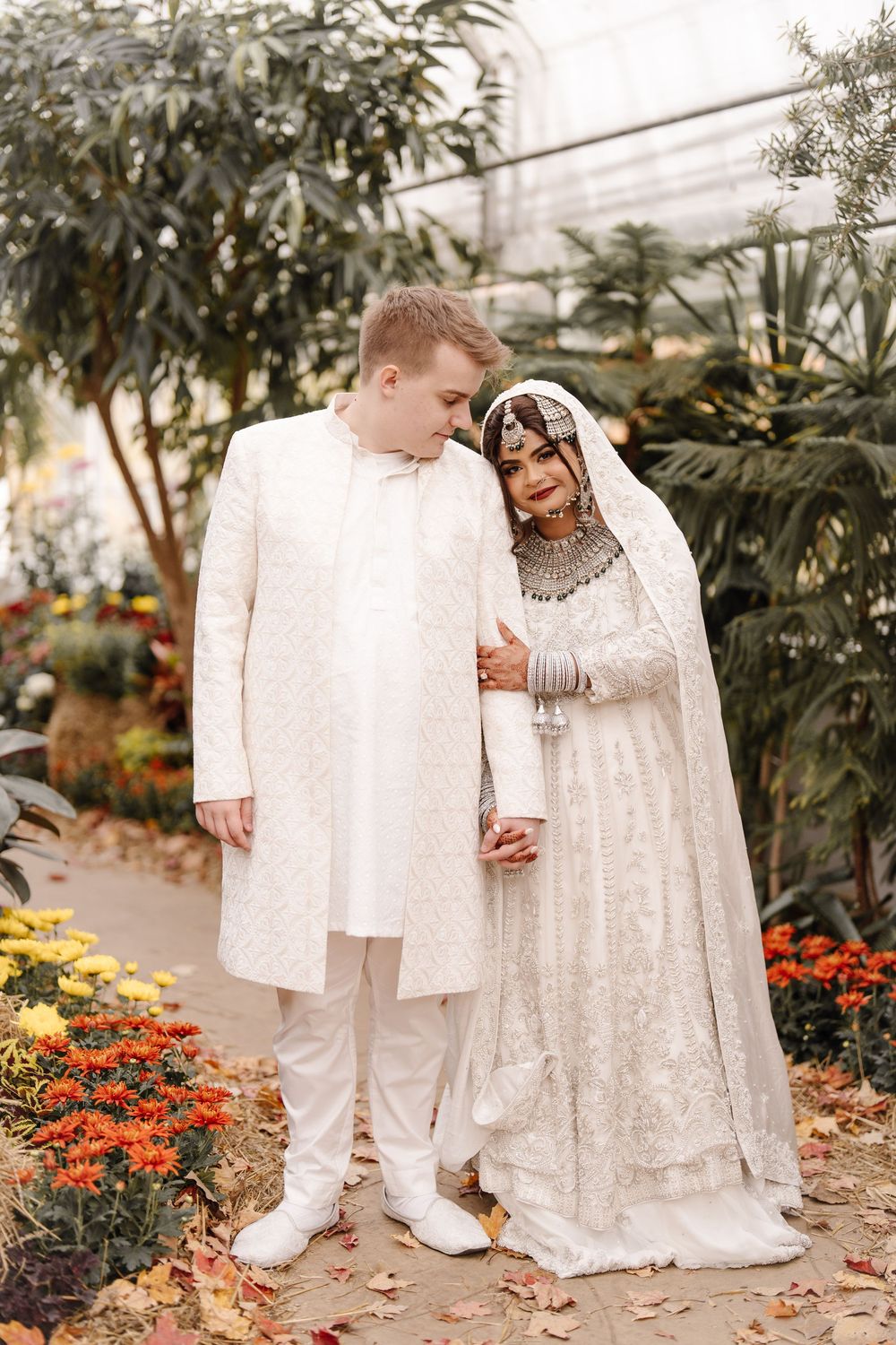 Interracial Muslim wedding couple in white traditional attire pose together among colorful flowers in greenhouse.